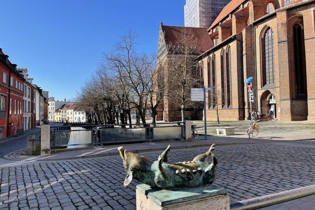 Ett torg med kullerstenar, en skulptur, en byggnad av rött tegel och en klarblå himmel.