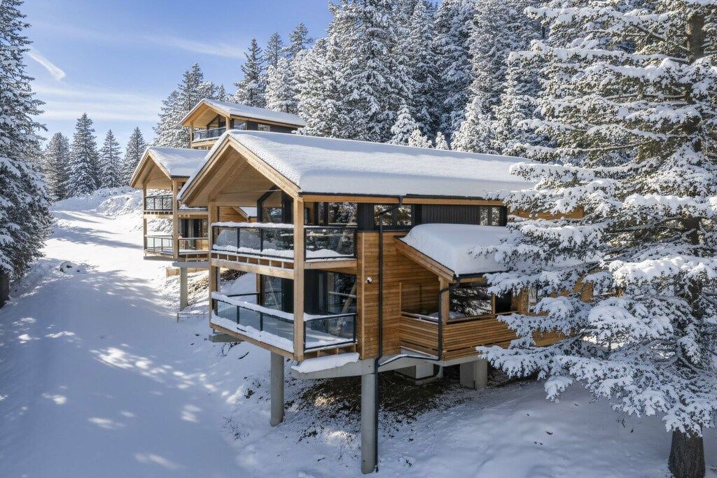 Une cabane en bois à plusieurs niveaux, dotée de balcons en verre, et entourée d’arbres couverts de neige.
