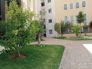 A courtyard with a paved walkway, a small tree, and a building with multiple windows and balconies. - A furnished apartment in Cité Ennasr, located on a private floor with no neighbors. (Ariana)