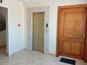 A hallway with a wooden door, an elevator, and a red emergency exit sign. - A furnished apartment in Cité Ennasr, located on a private floor with no neighbors. (Ariana)