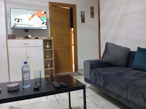 A living room with a sofa, a coffee table with a bottle and remote controls, a TV on the wall, and a door with a shelf above it. - A furnished apartment in Cité Ennasr, located on a private floor with no neighbors. (Ariana)