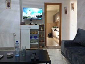 A living room with a television, a sofa, a coffee table with remote controls and a water bottle, and a kitchen visible through an open door. - A furnished apartment in Cité Ennasr, located on a private floor with no neighbors. (Ariana)