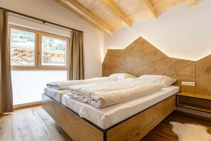 Ein Schlafzimmer mit einem Bettgestell aus Holz, einem Kopfteil, einem Fenster mit Vorhängen und Blick auf eine Holzterrasse.