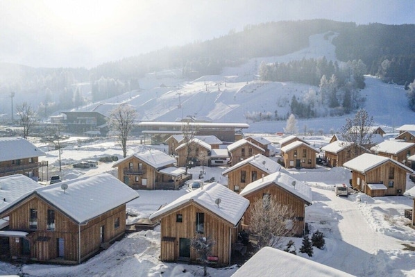 Un groupe de chalets en bois aux toits enneigés, entourés d’un paysage hivernal et avec un télésiège visible au loin.