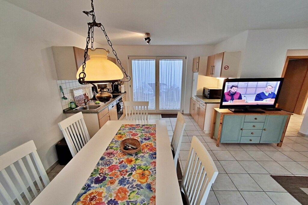 A dining area with a table set for four, a television on a stand, and a kitchen area with cabinets and a window.