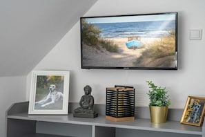 Una televisione a parete mostra una spiaggia, insieme a una fotografia incorniciata, una statua di Buddha, una lanterna nera e dorata e una pianta in vaso.