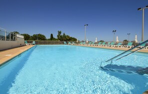Ein klares, blaues Schwimmbecken mit einer Metallleiter, umgeben von weißen Liegestühlen und Sonnenschirmen, unter blauem Himmel.
