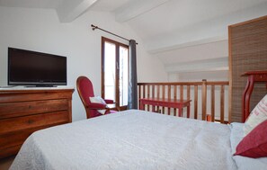 Ein Schlafzimmer mit einem Bett, einer Holzkommode, einem Fernseher, einem Sessel, einem Fenster mit Vorhängen und einem Holzlaufgeländer.