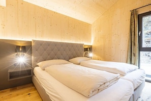 A bedroom with a wooden ceiling, a tufted headboard, two bedside tables with lamps, and a window with curtains. - Superior Chalet #23 with Sauna & Hot Tub (Turrach)