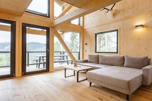 A wooden interior with a sofa, a table, and large windows offering a view of the outdoors. - Superior Chalet #23 with Sauna & Hot Tub (Turrach)