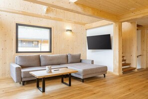 A modern interior with a wooden ceiling, a grey sofa, a wooden coffee table, a flat-screen TV mounted on the wall, and a staircase leading to an upper level. - Superior Chalet #23 with Sauna & Hot Tub (Turrach)