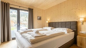 Quarto de hotel com cama grande, teto de madeira, vista por uma porta de vidro e uma pequena mesa de cabeceira.