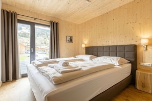 Ein Hotelzimmer mit einem großen Bett, einer Holzbalkendecke, Blick durch eine Glastür und einem kleinen Nachttisch.