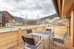 Eine Holzdielen-Terrasse mit Tisch und Stühlen, mit Blick auf eine gebirgige Landschaft und ein Club Hotel Gebäude.