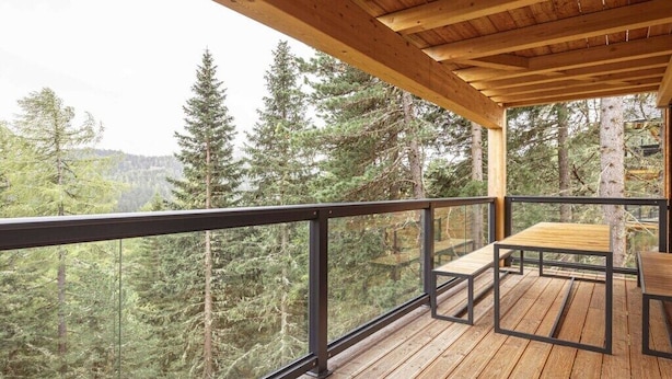Um deck de madeira com guarda-corpo de metal, uma mesa e bancos, com vista para uma área arborizada.