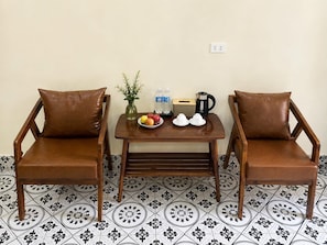 Two brown leather chairs with cushions, a small wooden table with a bowl of fruit, a water bottle, a kettle, and a vase with green plants. - Tam Coc Amana Boutique Hotel (Nam Hoa Lu)