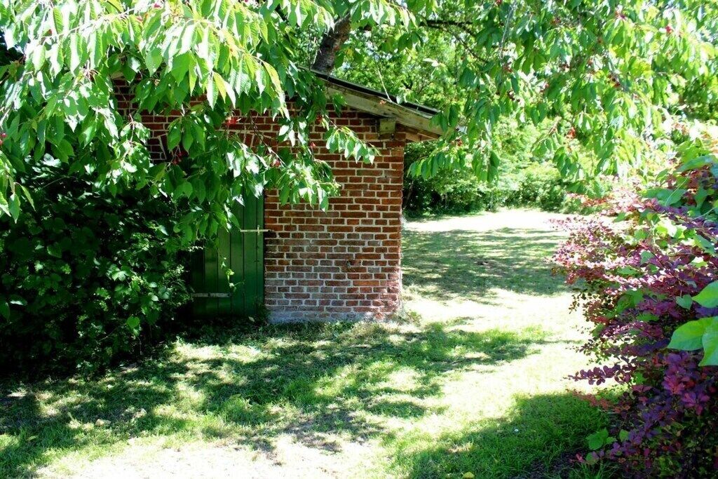 Edifício de tijolos com telhado inclinado, cercado por vegetação exuberante e um gramado.