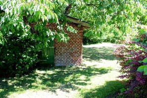 Edifício de tijolos com telhado inclinado, cercado por vegetação exuberante e um gramado.