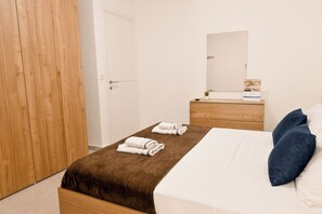 Een slaapkamer met een houten garderobe, een bed met wit beddengoed en een bruine deken, een houten nachtkastje met een spiegel, en een kleine potplant.