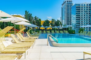 A swimming pool area with lounge chairs and umbrellas, surrounded by greenery and a tall building. - Unique Terrace in Midtown Pool (Miami)