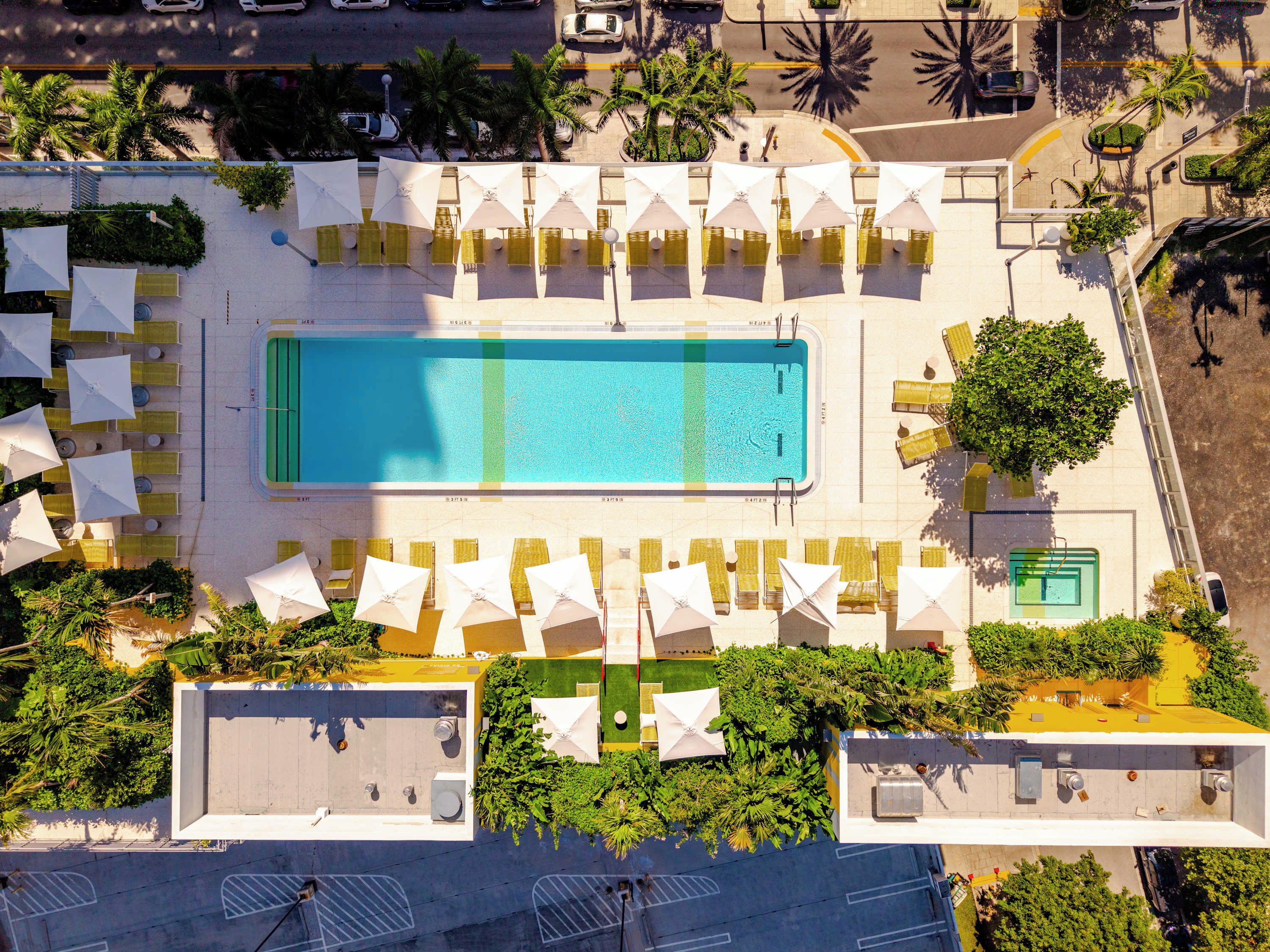 An aerial view of a hotel with a central swimming pool, multiple lounge areas with umbrellas, and surrounding greenery.