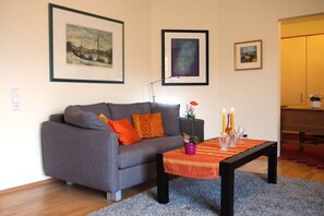 A living room with a grey sofa, orange cushions, a coffee table with candles, and framed artwork on the wall. - Holiday apartment with 1 bedroom (Bad Zwischenahn)