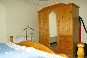 A bedroom with a wooden wardrobe, a bed with a blue and white bedspread, and a red vacuum cleaner. - Holiday on the North Sea coast in Pilsum (Krummhörn Pilsum)