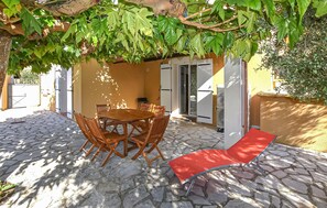 Eine Außenpatio mit Steinboden, einem Holztisch mit Stühlen, einem roten Liegestuhl und einer Pergola mit Grünpflanzen.