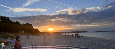 A beach with a sunset, a pier in the distance, and a striped beach umbrella.