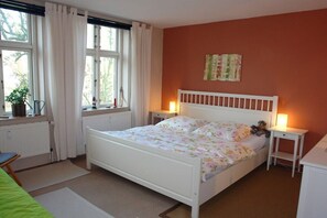 A bedroom with a bed, bedside tables, a radiator, a window with curtains, and a wall-mounted artwork. - Holiday apartment with 1 bedroom (Göhren)
