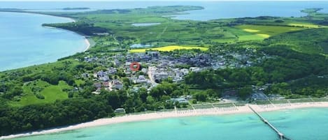 An aerial view of a coastal area with a beach, a pier, and a residential area with houses and greenery.