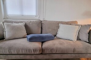 A beige sofa with four cushions, one blue, in a room with a window and white walls. - Thatched house "Alana" (Zierow)