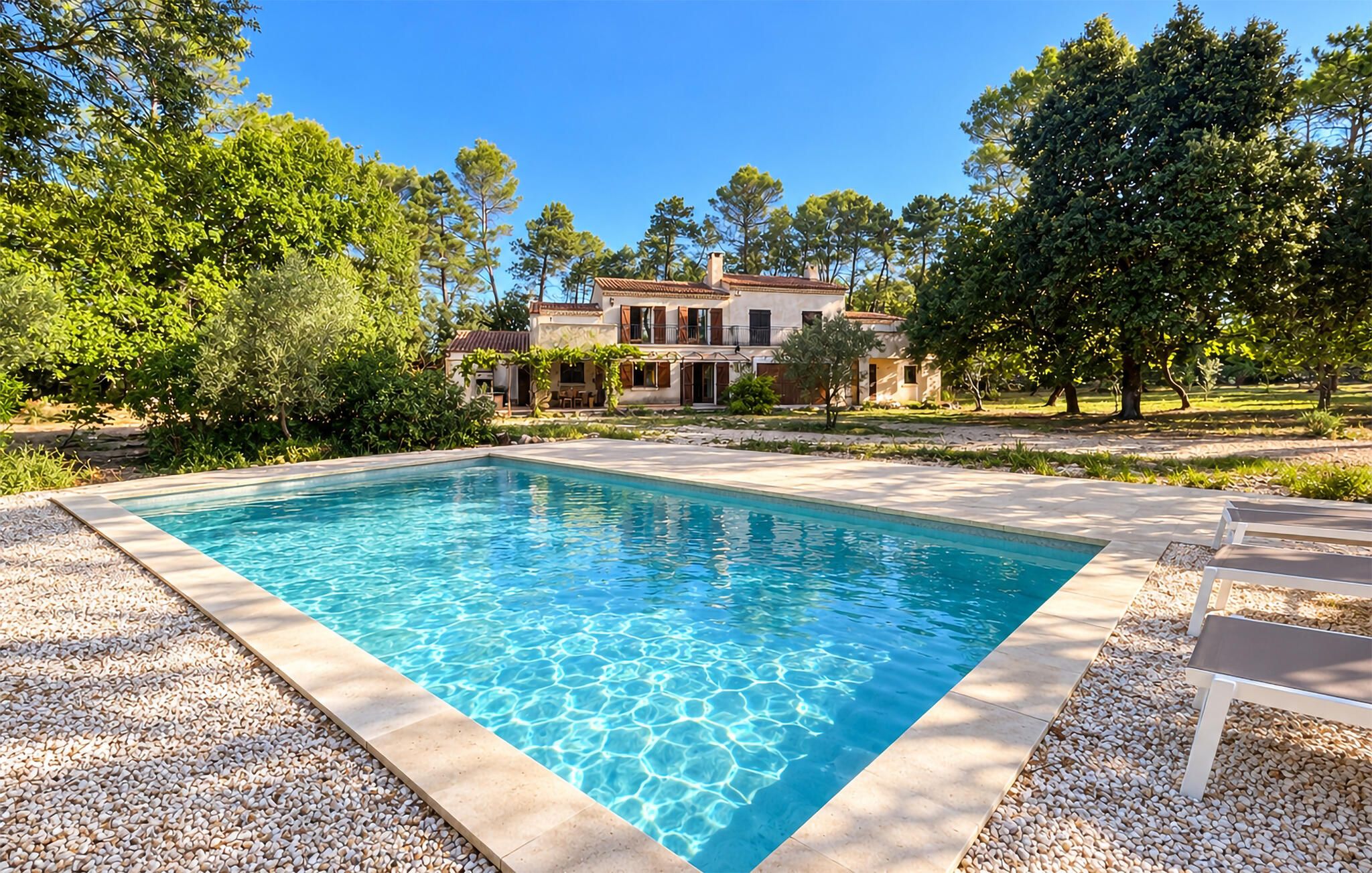 Una piscina rectangular con agua azul cristalina, rodeada por un borde de guijarros. Al fondo, un edificio grande con techo de tejas y varias ventanas.
