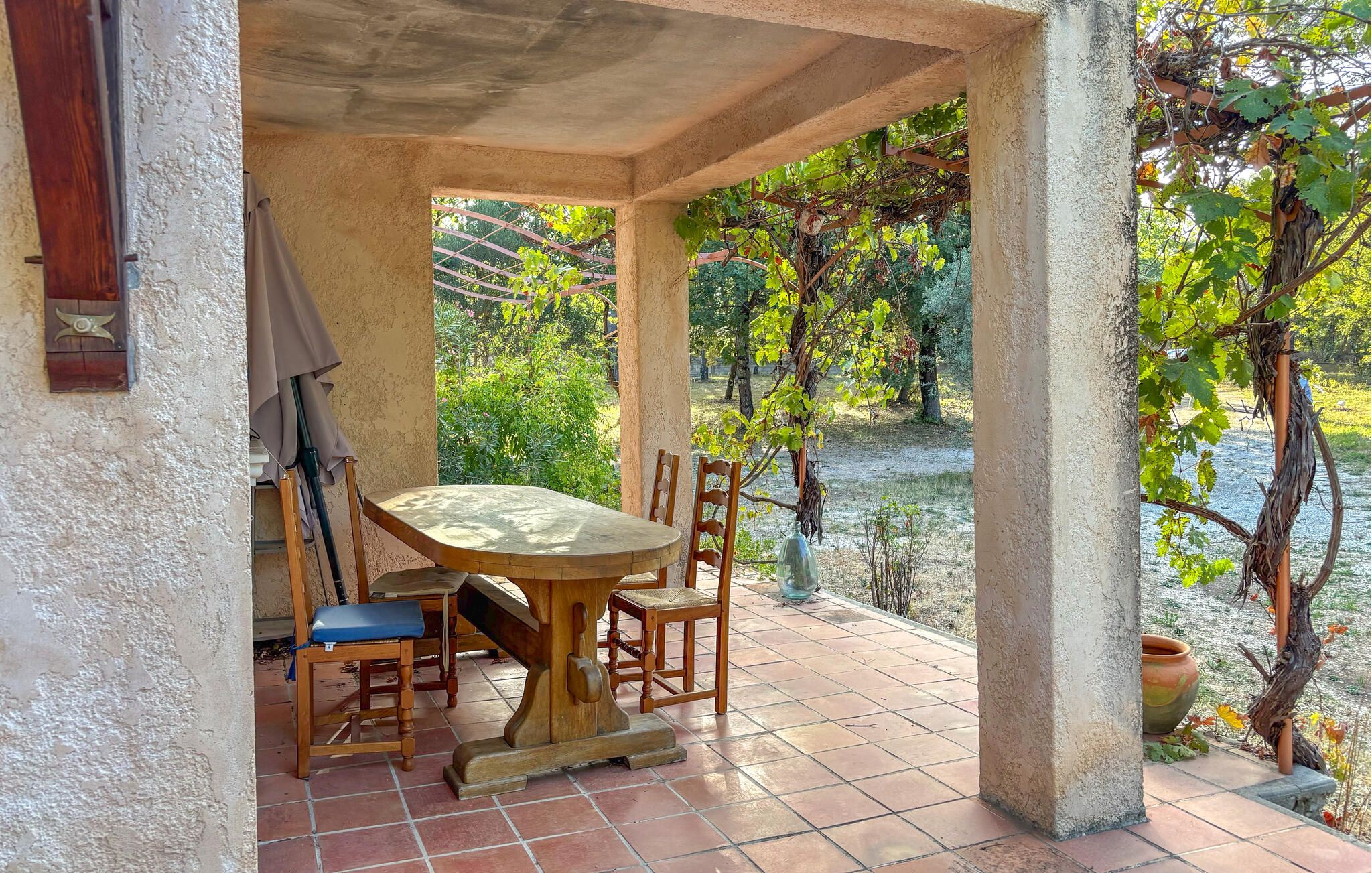 Une terrasse couverte avec une table et des chaises, offrant une vue sur la verdure et un plan d’eau.