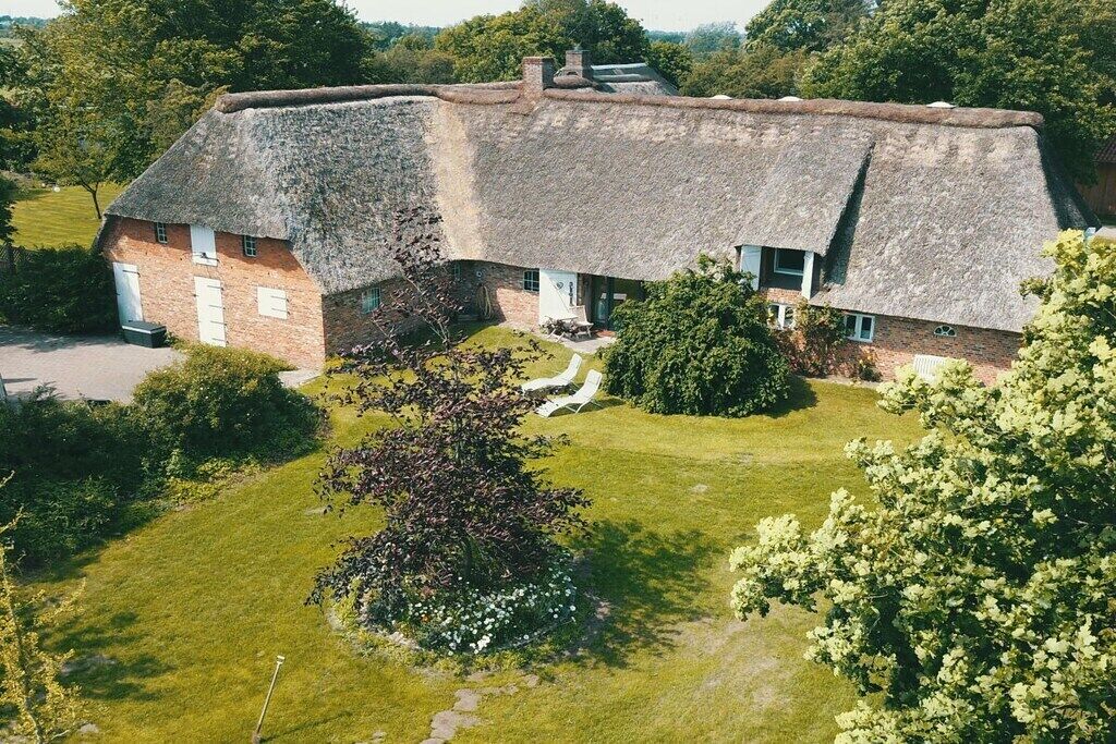 Un bâtiment à toit de chaume, avec une façade en briques, entouré d’une pelouse bien entretenue et d’arbres.