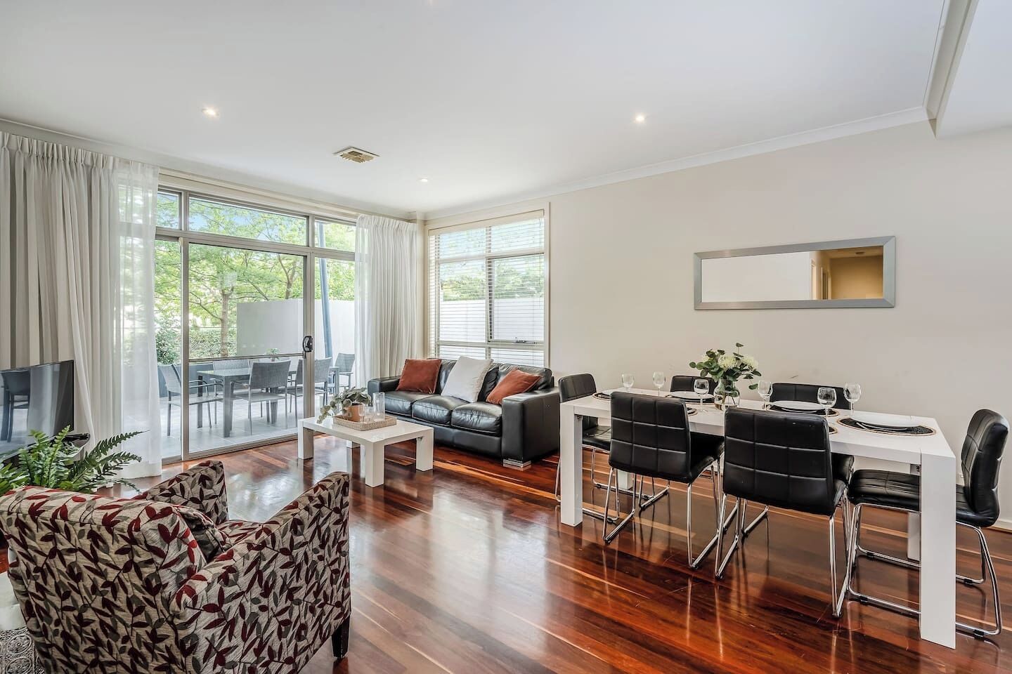 A spacious living area with a sofa, dining table, and chairs, large windows, and a flat-screen TV.