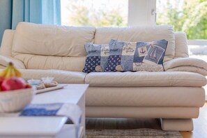 Ein beiges Sofa mit drei Zierkissen, einer Obstschale und einem Tisch mit einem Buch und einer Fernbedienung.