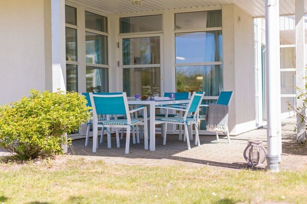 Een overdekt buitengebied met een tafel, stoelen, een kleine plant en een wand met ramen.