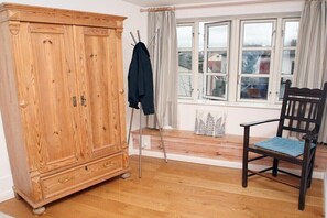 A room with a wooden wardrobe, a chair with a cushion, a window with curtains, and a coat rack with a jacket. - Holiday apartment with 1 bedroom (Flensburg)