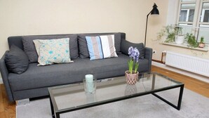 Ein modernes Wohnzimmer mit grauem Sofa, gemusterten Kissen, einem Glastisch, einer kleinen Vase mit Blumen, einer Stehlampe und einem Fenster mit Pflanzen.