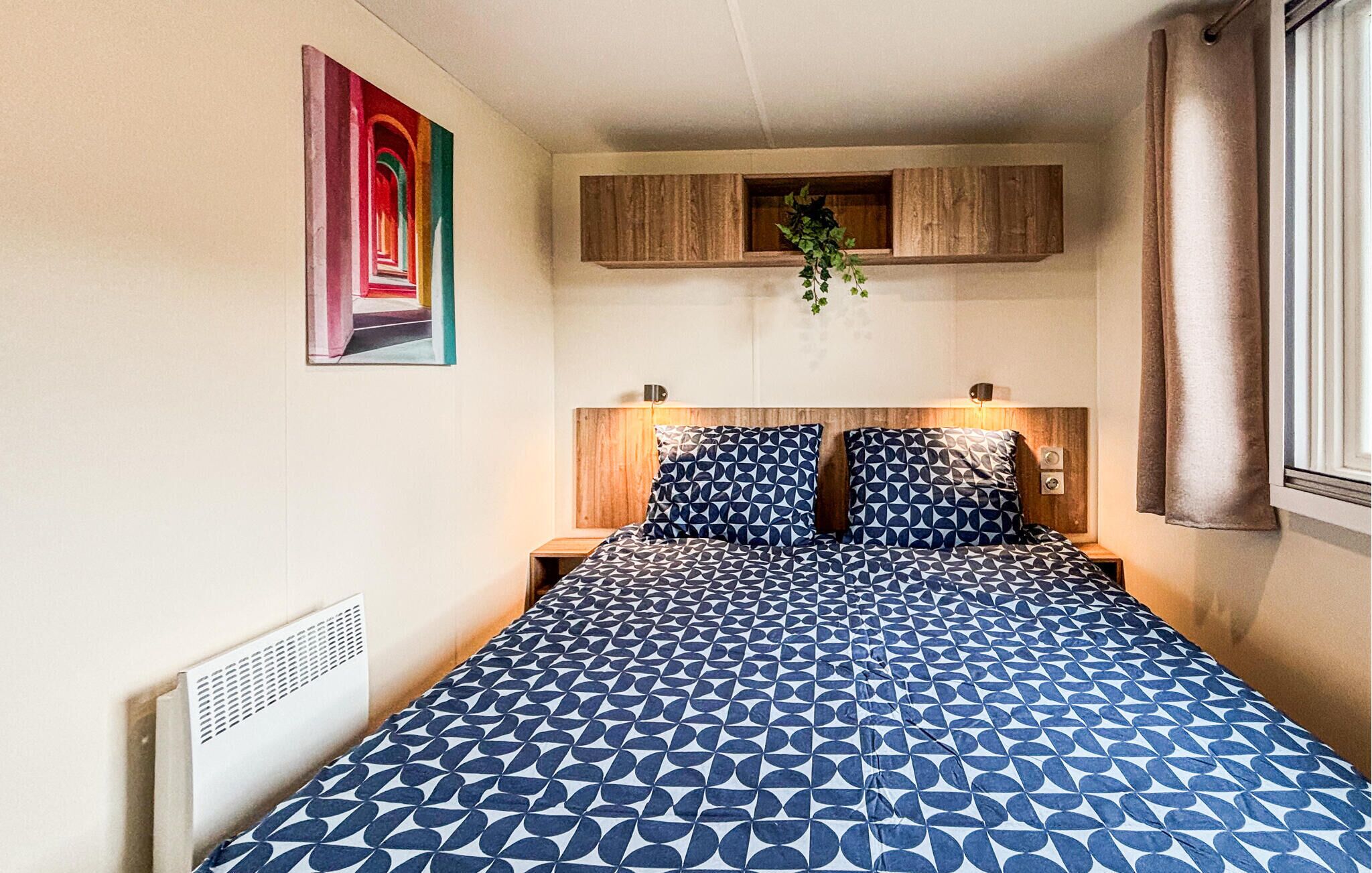 Une chambre à coucher avec une couvre-lit à motifs, une tête de lit dotée d’un éclairage intégré, une œuvre d’art accrochée au mur et une fenêtre avec des rideaux.