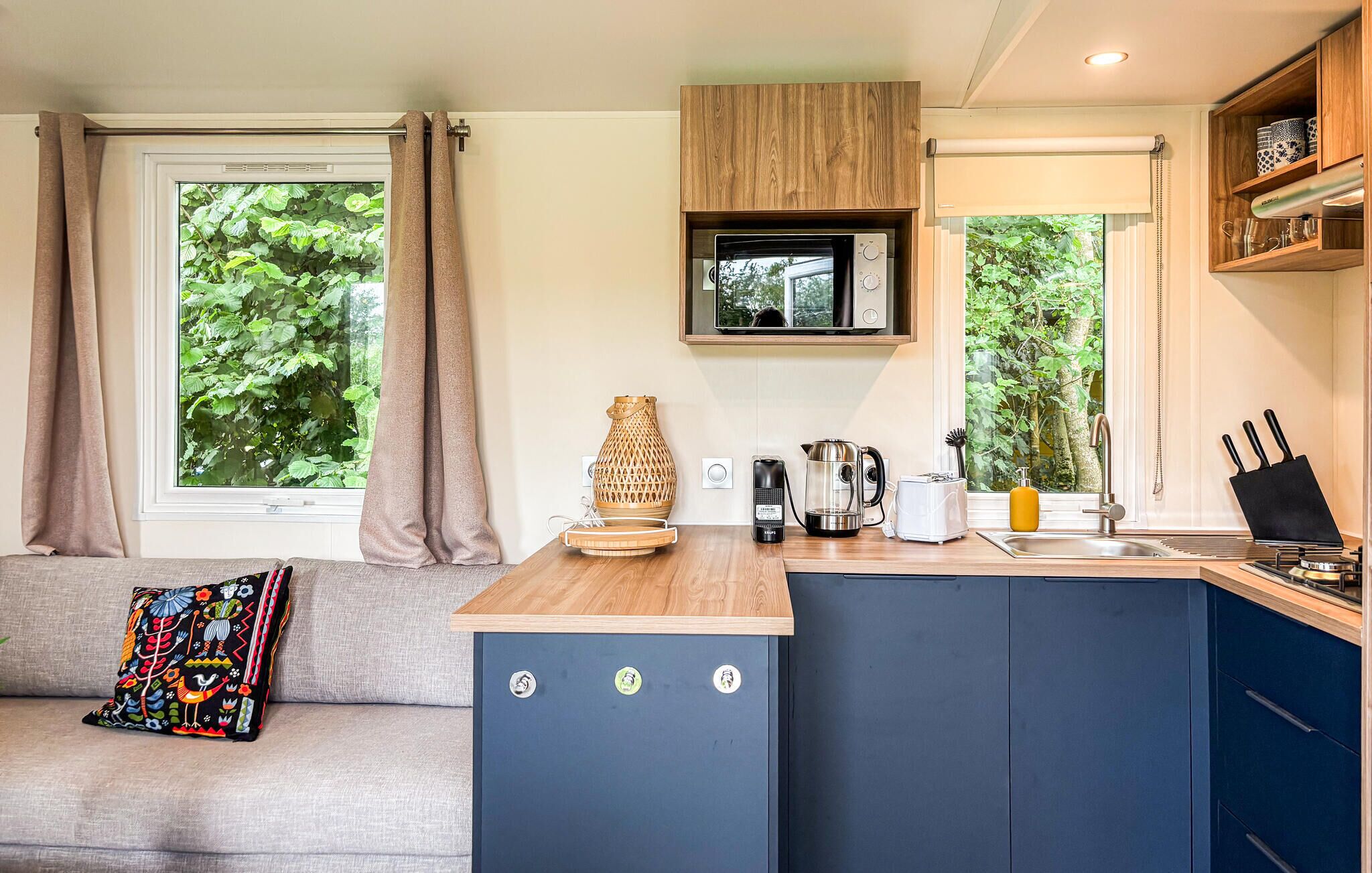 A compact kitchenette with a built-in microwave, coffee maker, and toaster. There is a window with a view of greenery outside, and a sofa with a colorful pillow.
