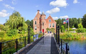 A wooden bridge leading to a historic brick building with a flag, surrounded by greenery and a calm water body. - Stunning home in Oude Pekela with sauna (Oude Pekela)