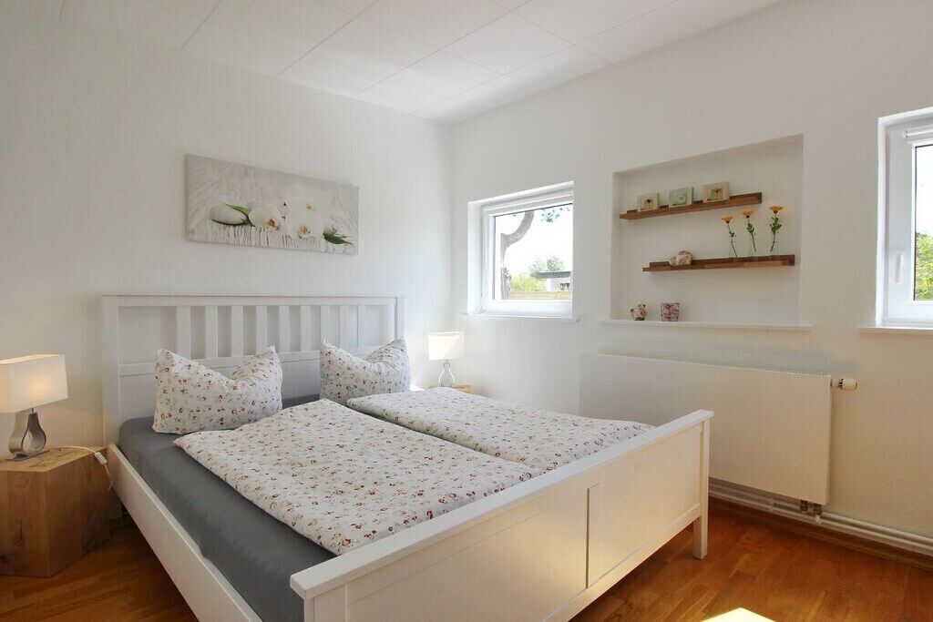 A bedroom with a white bed, a nightstand, a window, and a wall-mounted shelf with decorative items.