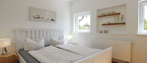 Un dormitorio con cama blanca, mesita de noche, ventana y estante montado en la pared con objetos decorativos.