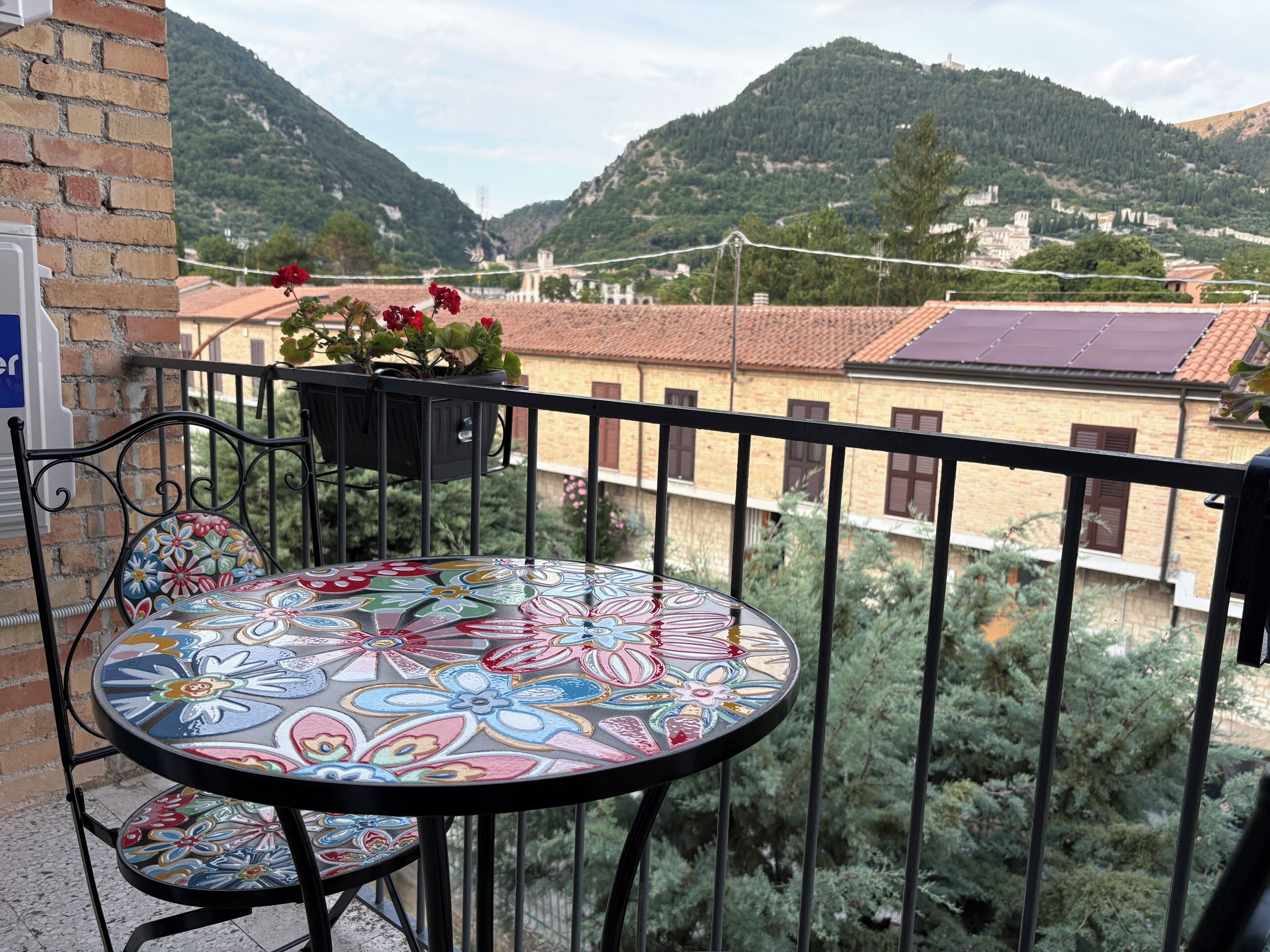 A balcony with a round table and chairs, a potted plant with flowers, and a view of a mountainous landscape with buildings.