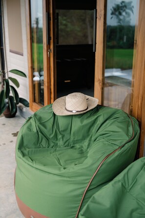 Un fauteuil en forme de sac de haricots vert, sur lequel est posé un chapeau de paille, est placé devant une porte vitrée donnant sur un espace intérieur sombre.