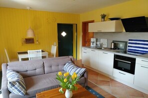 Ein Wohnzimmer mit einer Couch, einem Couchtisch mit Blumen, einem Essbereich mit Tisch und Stühlen sowie einer Küche mit weißen Schränken und Geräten.