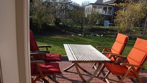 Eine Terrasse mit einem Holztisch und orangenen Stühlen, einem Ziegelboden und Blick auf ein Haus und einen Garten.