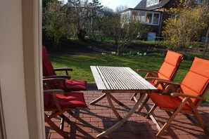 Une terrasse avec une table en bois et des chaises orange, un sol en briques et une vue sur une maison et un jardin.